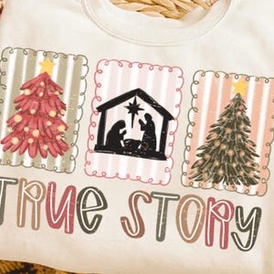May include: Cream-colored sweatshirt with a Christmas-themed design. The design includes three framed images: a red Christmas tree, a nativity scene, and a green Christmas tree. The text "TRUE STORY" is printed below the images.