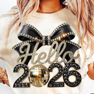 May include: Cream-colored t-shirt featuring a black bow embellished with rhinestones, the word "Hello" in a sparkling font, and the numbers "2026" with a disco ball in the center. The design is centered on the shirt.