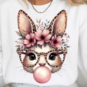 May include: White sweatshirt featuring a whimsical bunny face with large ears, wearing glasses and blowing a bubble. The bunny has a floral crown with pink flowers. The glasses and ears have a leopard print design. A fun, playful design.