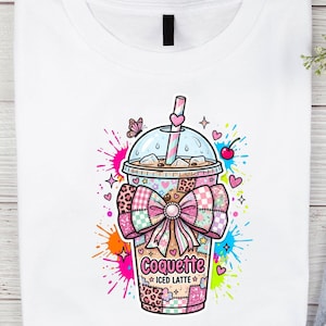 Op de afbeelding: Wit T-shirt met een kleurrijke afbeelding van een ijskoffie. Het ontwerp bevat een strik, hartjes en de woorden "Coquette Iced Latte". De afbeelding heeft een patchwork-ontwerp met roze, blauwe en oranje verfspatten.