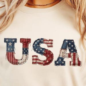 May include: Cream-colored t-shirt with the word "USA" in a patchwork design. The letters are in red, white, and blue, with stars and stripes patterns, evoking an American flag theme. The shirt is a casual wear item.