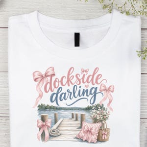 May include: A white t-shirt featuring a graphic print with the text "dockside darling" in pink and blue script. The design includes illustrations of a wooden dock over water, pink bows, a floral arrangement in a basket, and a pink ruffled pillow.