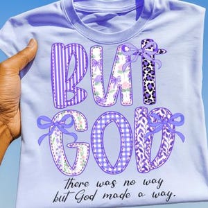 May include: A light purple t-shirt with the words "BUT GOD" in a patchwork design. The letters have various patterns, including stripes, floral, and leopard print, with purple bows. The text "There was no way but God made a way" is printed below.