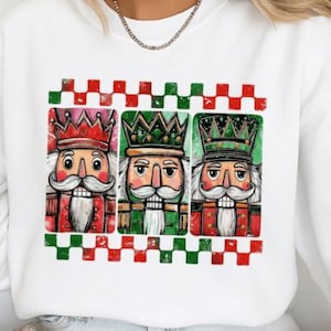 以下が含まれることがあります： 3体のくるみ割り人形のグラフィックが特徴の白いスウェットシャツ。各人形は異なる王冠と制服を着ており、赤と緑のチェック柄のボーダーが施されています。ホリデーシーズン向けのフェスティバルデザインです。