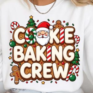 May include: White sweatshirt with a colorful graphic that reads "COOKIE BAKING CREW." The design features a gingerbread man, Santa Claus, candy canes, Christmas trees, and festive ornaments. The text is in a brown, cookie-like font.