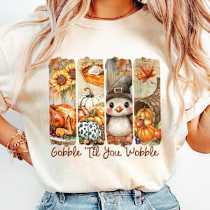 May include: Cream-colored t-shirt with a colorful fall-themed graphic. The graphic features sunflowers, a pumpkin pie, pumpkins, a turkey, a penguin in a witch hat, and a cornucopia. The text "Gobble 'Til You Wobble" is printed below the graphic.