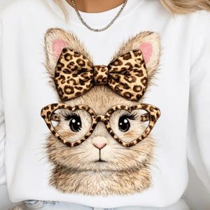 May include: White sweatshirt featuring a detailed illustration of a bunny face. The bunny wears heart-shaped glasses and a bow, both with a leopard print design. The bunny's fur is light brown, with pink accents on the ears.