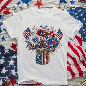 May include: A white short-sleeve t-shirt featuring a central graphic of a mason jar decorated with American flag patterns, filled with a bouquet of sparkling red, white, and blue jeweled flowers and two small American flags on sticks.