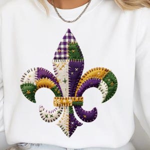 May include: White sweatshirt featuring a colorful fleur-de-lis design. The fleur-de-lis is made of patchwork in green, purple, gold, and white. The design is centered on the front of the shirt.