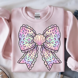 May include: A pale pink sweatshirt with a large bow design. The bow features a leopard print pattern in pastel shades, with a seashell in the centre. The sweatshirt is made of a soft material.