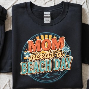 May include: Black sweatshirt with a circular graphic that reads "MOM needs a BEACH DAY" in orange, blue, and yellow. The design includes palm trees and a wave. The Gildan tag is visible.