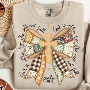 May include: Beige sweatshirt with a large bow design featuring a cross and floral and plaid patterns. The text "She will not fall, God is within her" is written around the bow. The scripture reference Psalm 46:5 is also included.