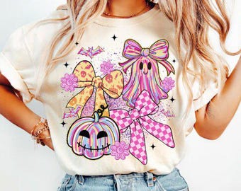 May include: Cream-colored t-shirt featuring a colorful Halloween-themed graphic. The design includes a ghost, pumpkin, and bows in pink, yellow, and checkered patterns. The graphic is accented with stars and floral elements.