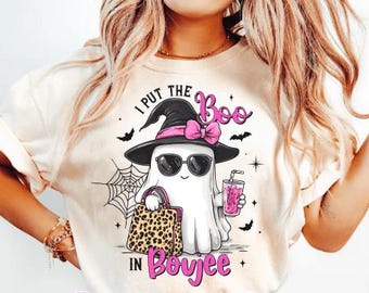 May include: Cream-colored t-shirt featuring a cartoon ghost wearing sunglasses, a witch hat with a pink bow, and holding a leopard print bag and a pink drink. The text reads "I PUT THE Boo IN Boujee". Bats and a spiderweb are also featured.
