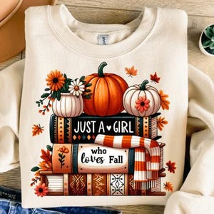 Peut inclure: Sweat-shirt crème avec un motif sur le thème de l'automne. Le motif présente des citrouilles, des fleurs et le texte "Just a Girl who loves Fall" sur des livres empilés. Le motif comprend des couleurs orange, blanc et marron.