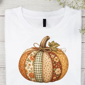 May include: A white t-shirt featuring a central graphic of a pumpkin composed of various fabric-like patches. The pumpkin displays orange, green, and cream floral and checkered patterns, tied with a thin brown twine bow at the top stem.