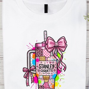 Puede incluir: Camiseta blanca con un gráfico colorido de un vaso Stanley Coquette rosa con pajita. El vaso tiene un diseño de patchwork con estampado de leopardo y el texto "Stanley Coquette". Un gran lazo rosa y salpicaduras de pintura coloridas completan el diseño.