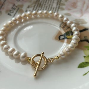 May include: A white pearl bracelet with a gold toggle clasp. The bracelet is on a white background with a floral pattern.