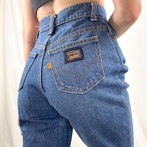 May include: High-waisted blue denim jeans featuring a dark blue rectangular patch on the back pocket with orange Levi Strauss & Co. text and logo. The jeans include an orange tab on the side of the pocket, belt loops, and visible stitching.