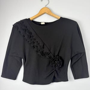May include: Black long-sleeve top with a round neckline and a decorative ruffle detail. The top features a floral embellishment at the waist and is displayed on a wooden hanger against a neutral background. A classic and versatile garment.
