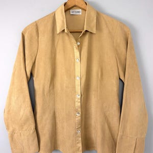 Puede incluir: Una camisa abotonada de color beige con mangas largas, cuello y puños abotonados. La camisa está colgada de una percha de madera. La etiqueta de la camisa dice "SATSUMA".