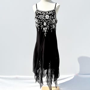 Puede incluir: Vestido negro con bordado floral blanco en el corpiño y el dobladillo. El vestido tiene tirantes finos y un dobladillo asimétrico. El vestido se exhibe en un maniquí sobre un fondo blanco.