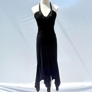 Puede incluir: Vestido negro con cuello halter, corpiño entallado y bajo asimétrico. El vestido presenta un adorno de cuentas en el escote y se exhibe en un maniquí. El vestido está confeccionado con una tela suave y fluida.