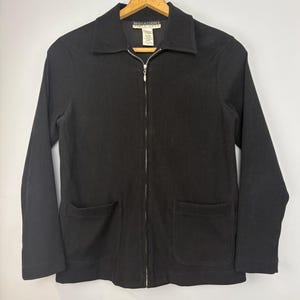 Puede incluir: Chaqueta negra con cremallera, dos bolsillos delanteros y cuello. La chaqueta está colgada en una percha de madera. El nombre de la marca "NOTATIONS" es visible en la etiqueta.