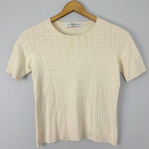 Könnte beinhalten: Cremefarbener Kurzarm-Zopfstrickpullover, der an einem Holzbügel hängt. Der Pullover hat einen Rundhalsausschnitt und ein strukturiertes Zopfmuster. Das Etikett lautet "sutton studio".