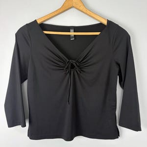 Puede incluir: Top negro con escote en V y mangas tres cuartos. La parte superior presenta un detalle fruncido en el busto con cierre de lazo. La prenda está colgada en una percha de madera sobre un fondo liso.