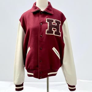 以下が含まれることがあります： 胸に「H」の文字が入った、マルーンとクリーム色のスタジアムジャンパー。クリーム色の袖、ボタン留めのフロント、ストライプの袖口と裾が特徴です。マネキンに着せられています。