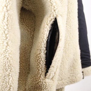 Pode incluir: Close-up de uma jaqueta de l&atilde; de cor creme com um z&iacute;per preto e um bolso preto. A l&atilde; &eacute; macia e fofa.