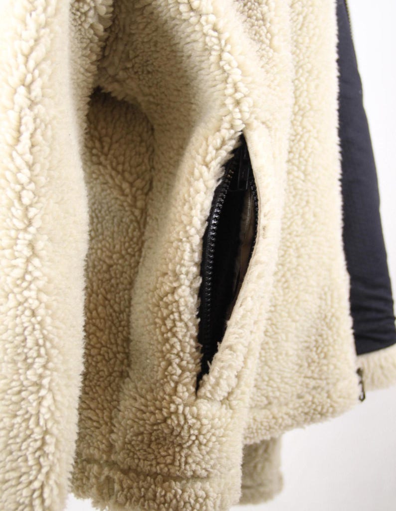 Pode incluir: Close-up de uma jaqueta de l&atilde; de cor creme com um z&iacute;per preto e um bolso preto. A l&atilde; &eacute; macia e fofa.