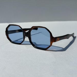 May include: Brown and black octagonal sunglasses with light blue lenses. The sunglasses have a tortoiseshell pattern on the frames and arms. The lenses are a light blue color, and the sunglasses are set against a white background.