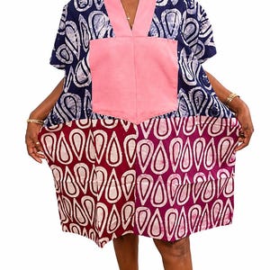 May include: A knee-length tunic dress featuring a V-neckline with a solid pink collar and a large central pink pocket. The upper section has a navy blue and white teardrop pattern, while the lower section features a maroon and white teardrop print.