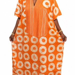 May include: A floor-length orange kaftan dress featuring a repeating pattern of large, light beige circles. The garment has short sleeves, a V-neckline, and a central vertical panel decorated with thin, white, parallel pinstripes running down the front.