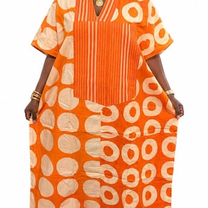 May include: A vibrant orange kaftan dress featuring a repeating pattern of large, light beige circles. The garment includes a V-neckline with a central panel of vertical white stripes, short sleeves, and a loose, flowing silhouette that reaches the floor.