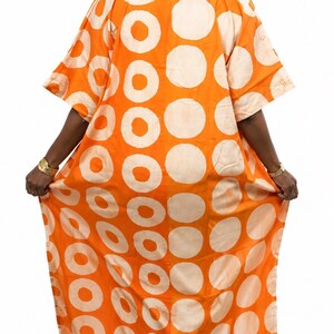 May include: A floor-length orange kaftan dress featuring a bold pattern of large, light cream-colored circles. The garment has short, wide sleeves, a high collar, and a loose, flowing silhouette that extends to the floor with a straight hemline.
