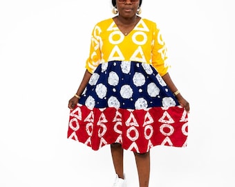May include: A knee-length dress featuring three horizontal color-blocked sections. The top section is yellow with white triangle patterns, the middle is navy blue with white circular motifs, and the bottom is red with white triangle patterns.