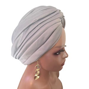 Peut inclure: Un turban gris clair avec des plis verticaux est porté sur une tête de mannequin. Des boucles d'oreilles dorées avec un motif circulaire superposé pendent de l'oreille. Le fond est blanc uni.