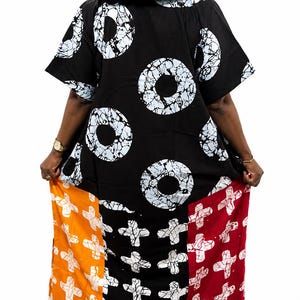 Pu&ograve; includere: Un abito caftano nero dal taglio ampio con un motivo batik circolare bianco sulla parte superiore e un pannello inferiore diviso in tre segmenti verticali di colore arancione, nero e rosso, decorati con un motivo a croce bianca.