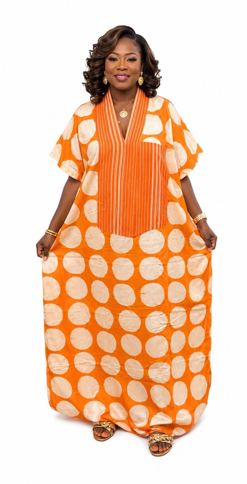 May include: A floor-length orange kaftan dress featuring a large white polka dot pattern throughout. The garment has short sleeves, a V-neckline, and a central vertical panel with thin white stripes that extends from the neckline to the mid-chest.