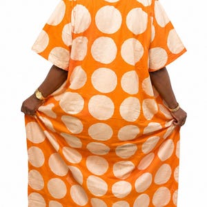 May include: A floor-length kaftan dress featuring a vibrant orange base with large, repeating cream-colored polka dots. The garment has short sleeves, a collared neckline with horizontal striped detailing, and a loose, flowing silhouette.
