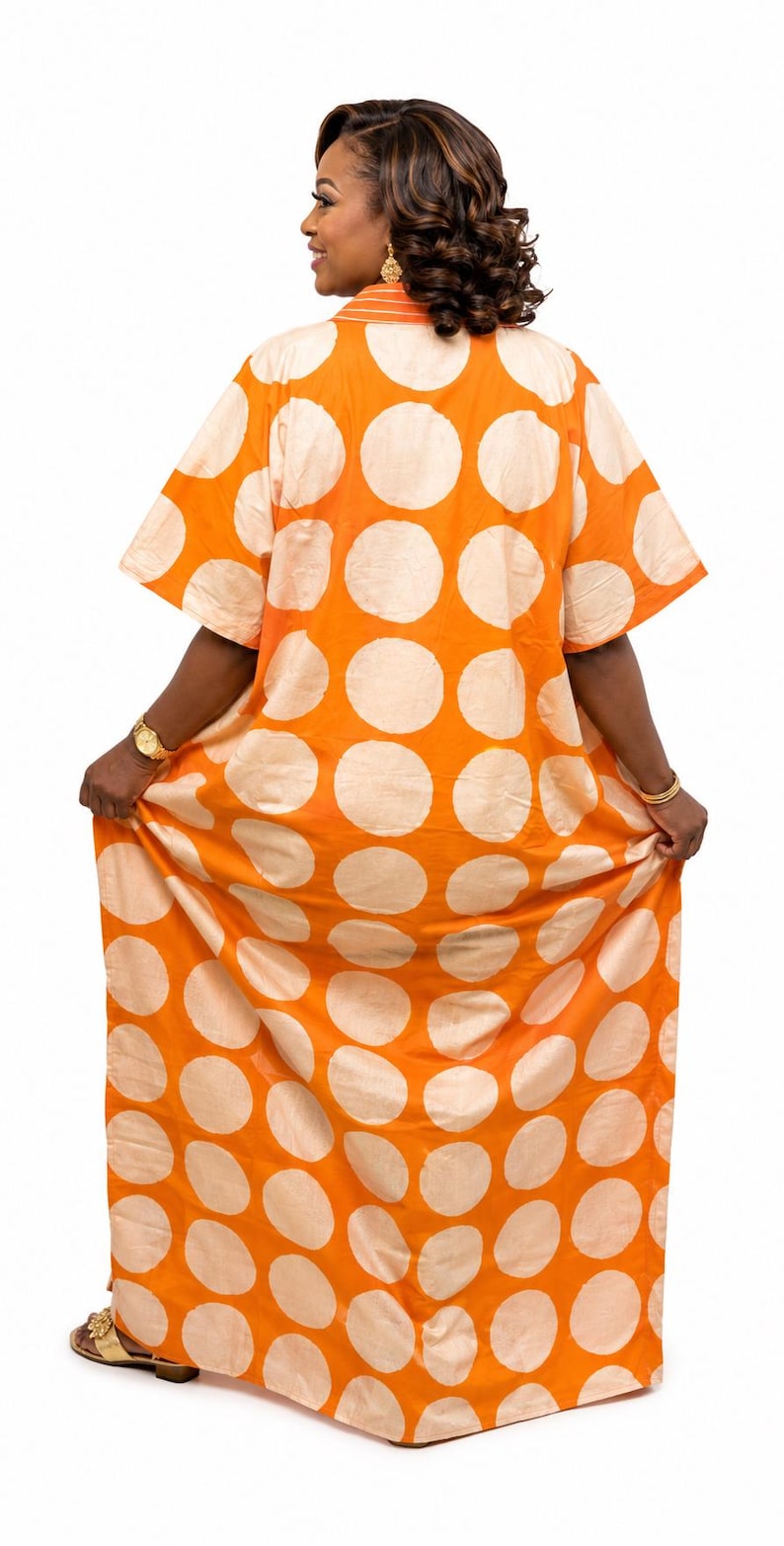 May include: A floor-length kaftan dress featuring a vibrant orange base with large, repeating cream-colored polka dots. The garment has short sleeves, a collared neckline with horizontal striped detailing, and a loose, flowing silhouette.