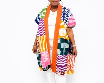 May include: A knee-length kimono-style jacket featuring a vibrant patchwork pattern of geometric and wavy shapes in green, purple, pink, yellow, and blue. The garment has a solid orange trim along the front opening and short, wide sleeves.