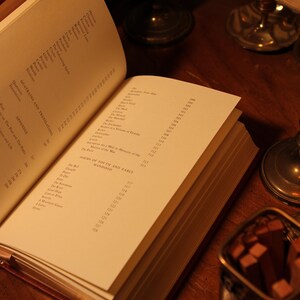 May include: An open book with pages displaying text, including "QUATRAINS AND TRANSLATIONS" and "POEMS OF YOUTH AND EARLY MANHOOD." The book is on a wooden surface, with a small metal container and candle holders in the background.