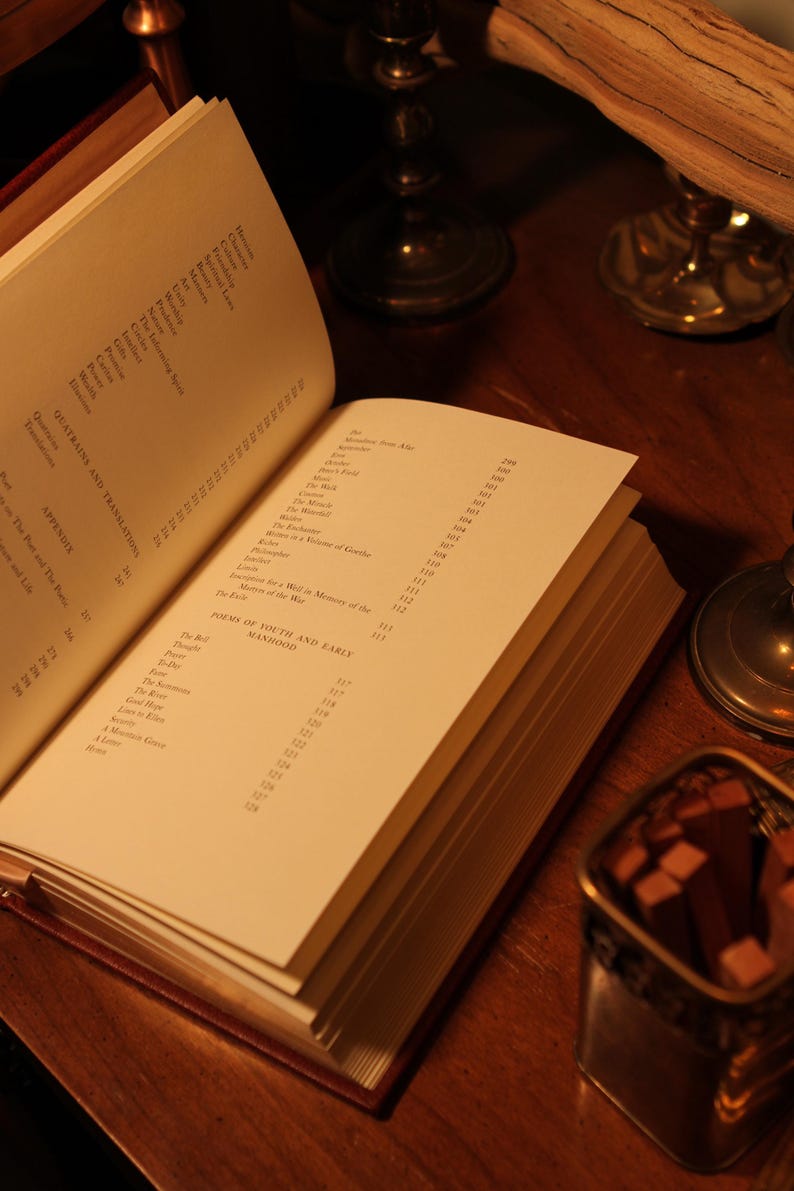May include: An open book with pages displaying text, including "QUATRAINS AND TRANSLATIONS" and "POEMS OF YOUTH AND EARLY MANHOOD." The book is on a wooden surface, with a small metal container and candle holders in the background.
