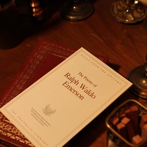 May include: A still life featuring a copy of "The Poems of Ralph Waldo Emerson" from The Franklin Library, resting atop a burgundy leather-bound book. The book's title is in a serif font. The scene is set on a dark wooden surface, with antique decor in the background.