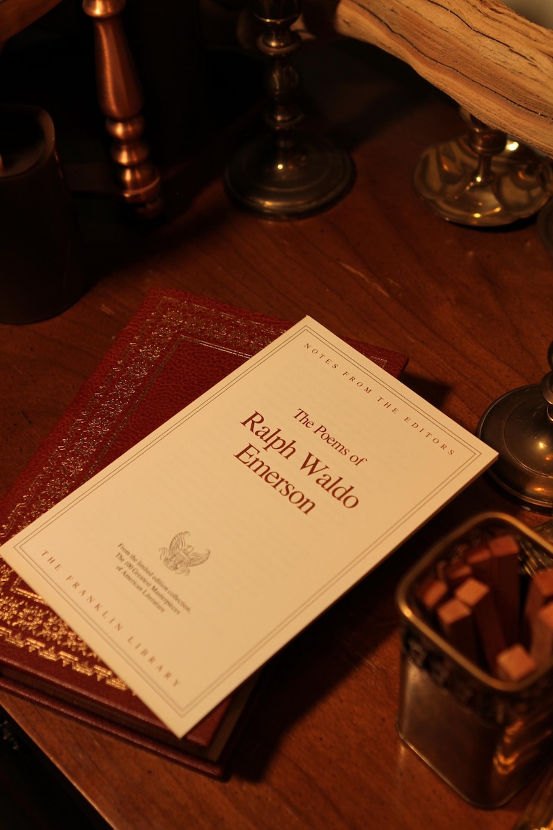May include: A still life featuring a copy of "The Poems of Ralph Waldo Emerson" from The Franklin Library, resting atop a burgundy leather-bound book. The book's title is in a serif font. The scene is set on a dark wooden surface, with antique decor in the background.