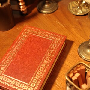 May include: A red leather-bound book with gold decorative trim. The book is on a wooden surface, alongside a candle, brass candlesticks, and a small container of matches. The book's cover has a textured appearance.